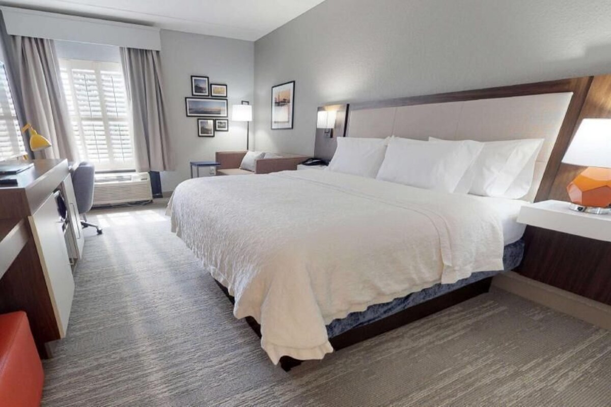 Habitación de hotel con una cama grande, un escritorio con ordenador, una silla, una lámpara de escritorio y un ventanal con cortinas.