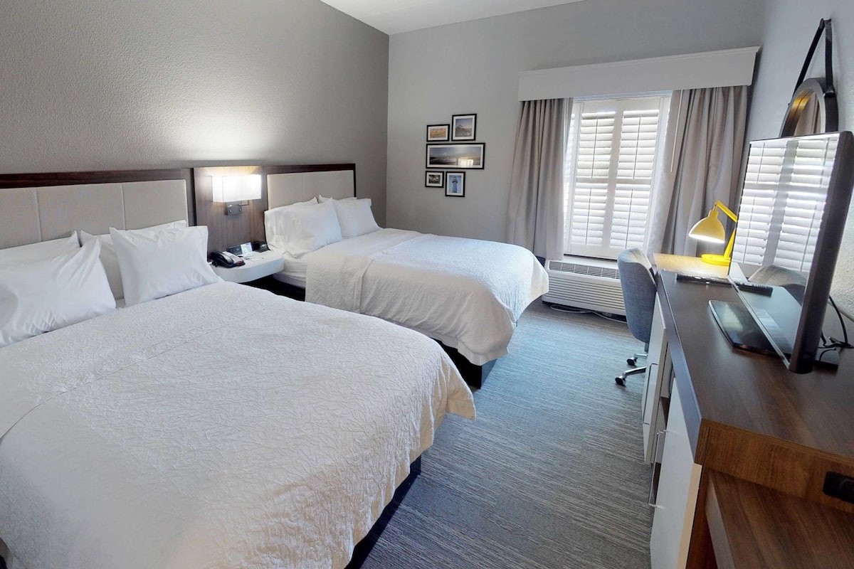 A hotel room with two beds, a desk with a chair, a desk lamp, a flat-screen TV, and a window with curtains.