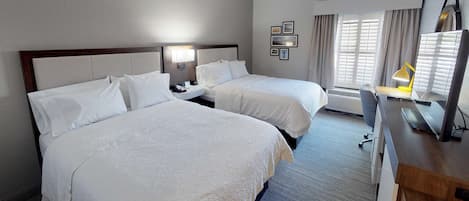 Habitación de hotel con dos camas, un escritorio con silla, una lámpara de escritorio, un televisor de pantalla plana y una ventana con cortinas.
