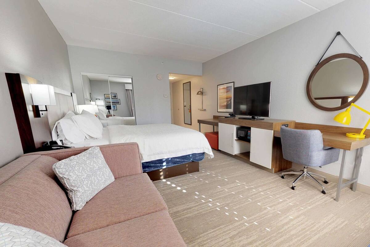 A hotel room with a bed, a desk with a computer, a chair, a TV, a mirror, and a lamp.