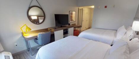 A hotel room with two beds, a desk with a computer and a TV, a mirror, and a small window.