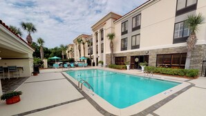 A pool area with lounge chairs and umbrellas, a stone wall, and a building with multiple windows and a tiled roof. - Hampton Inn St. Simons Island (St. Simons Island)