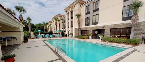 A pool area with lounge chairs and umbrellas, a stone wall, and a building with multiple windows and a tiled roof.