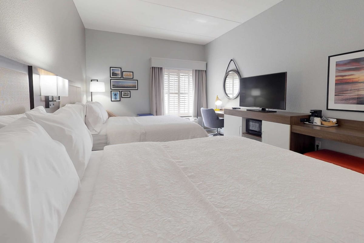 A hotel room with two beds, a desk with a television, a chair, and a window with curtains.