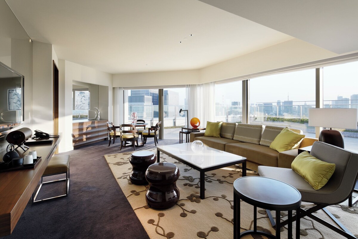 A modern living room with a sofa, coffee table, and chairs, large windows with a city view, and a desk with a lamp.