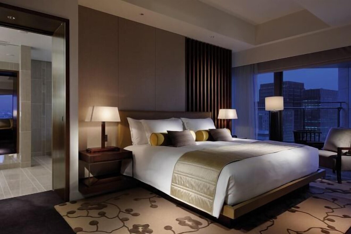A modern hotel room with a large bed, two bedside lamps, a nightstand, and a view of the city at dusk.