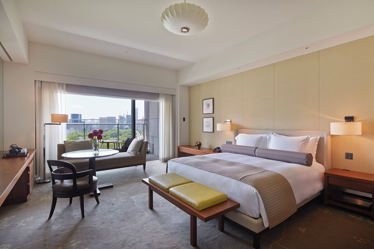 A hotel room with a bed, a desk, a chair, a bench, a bedside table, and a view of the city.