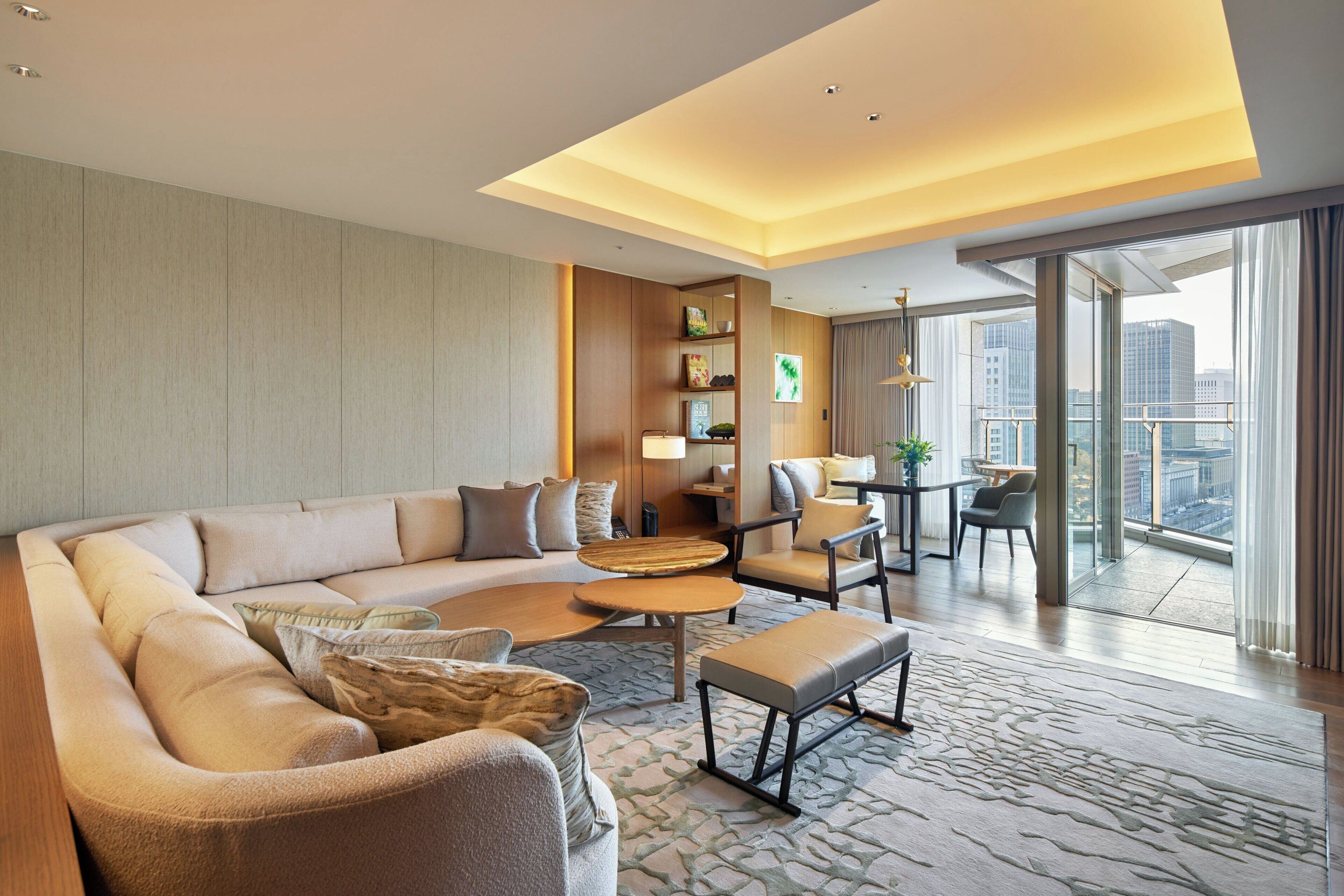 A modern hotel room with a sectional sofa, a small coffee table, a dining area with a table and chairs, a bookshelf, and a large window offering a city view.