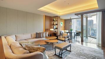 A modern hotel room with a sectional sofa, a small coffee table, a dining area with a table and chairs, a bookshelf, and a large window offering a city view.