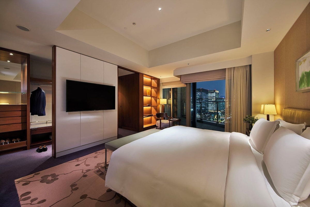 A hotel room with a large bed, a TV mounted on the wall, a wardrobe, a seating area with a table and chairs, and a view of the cityscape through the window.