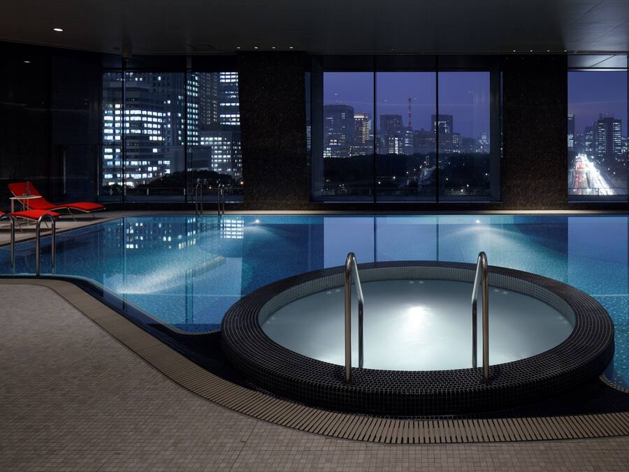 A modern indoor swimming pool with a hot tub, red lounge chairs, and a cityscape view through large windows.