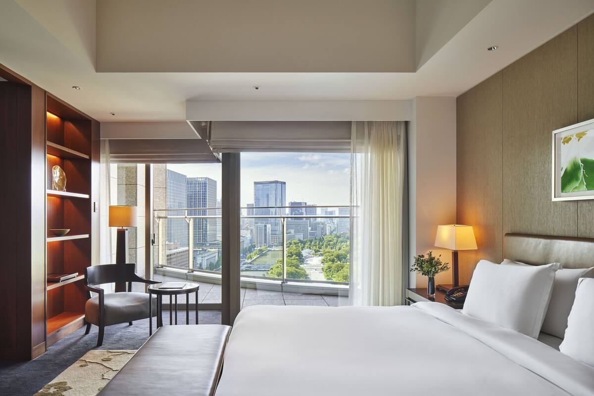 A hotel room with a large bed, a desk, a chair, a bookshelf, and a view of a cityscape through large windows.