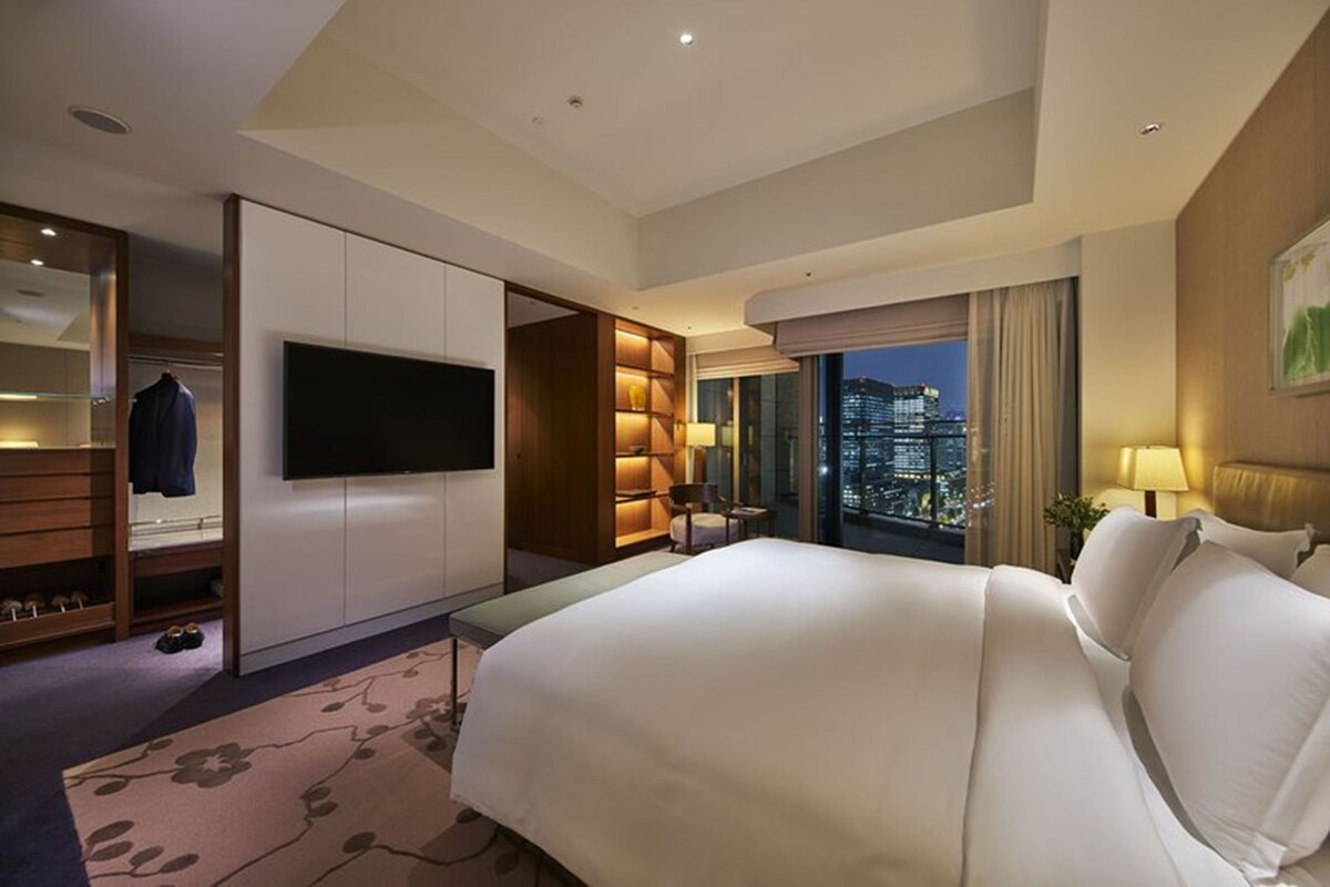 A hotel room with a large bed, a TV, a seating area with a view of the city, and a wardrobe.