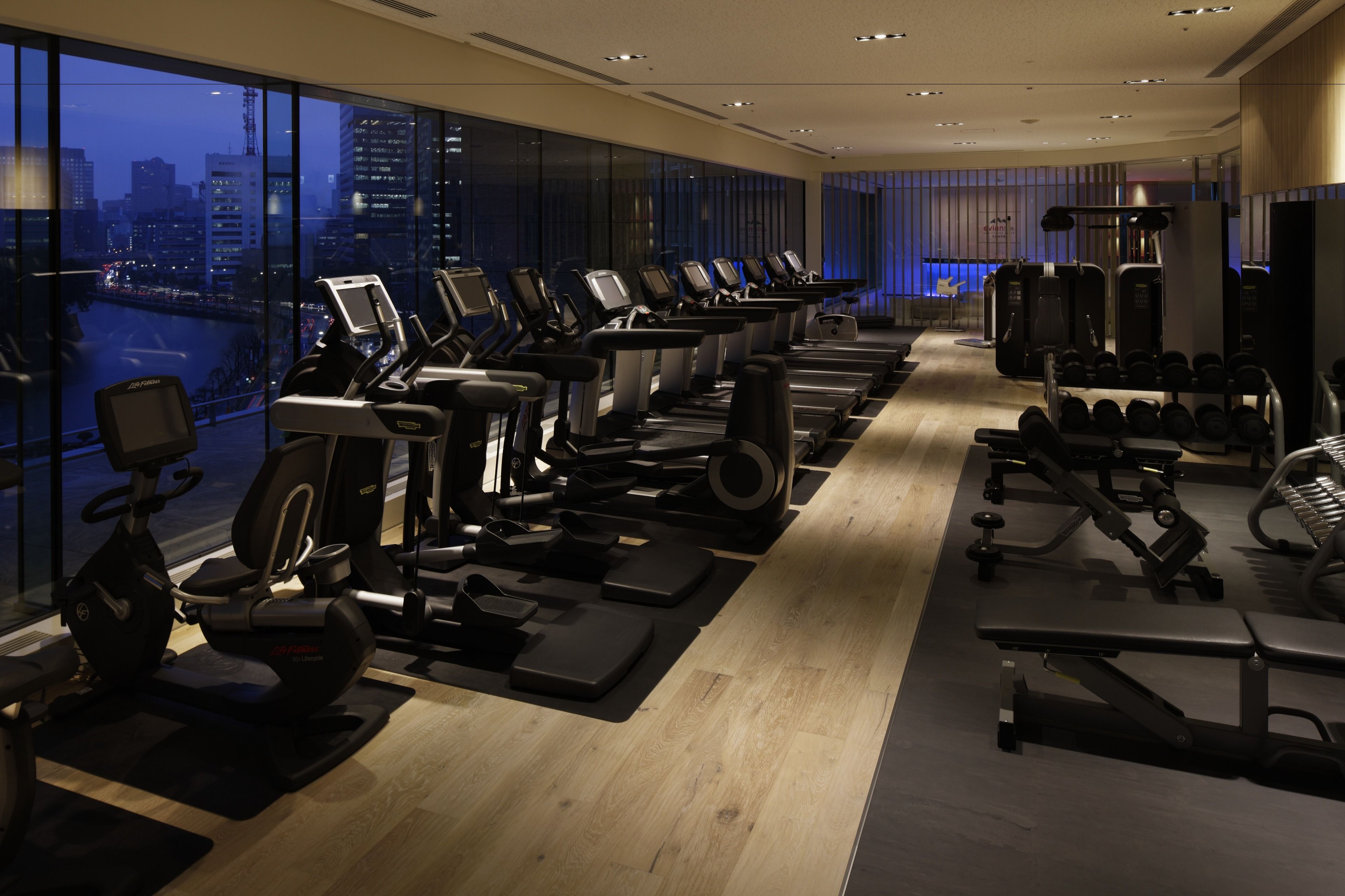 Une salle de sport moderne équipée de divers appareils d’exercice, de grandes fenêtres offrant une vue sur la ville et d’un plancher en bois.