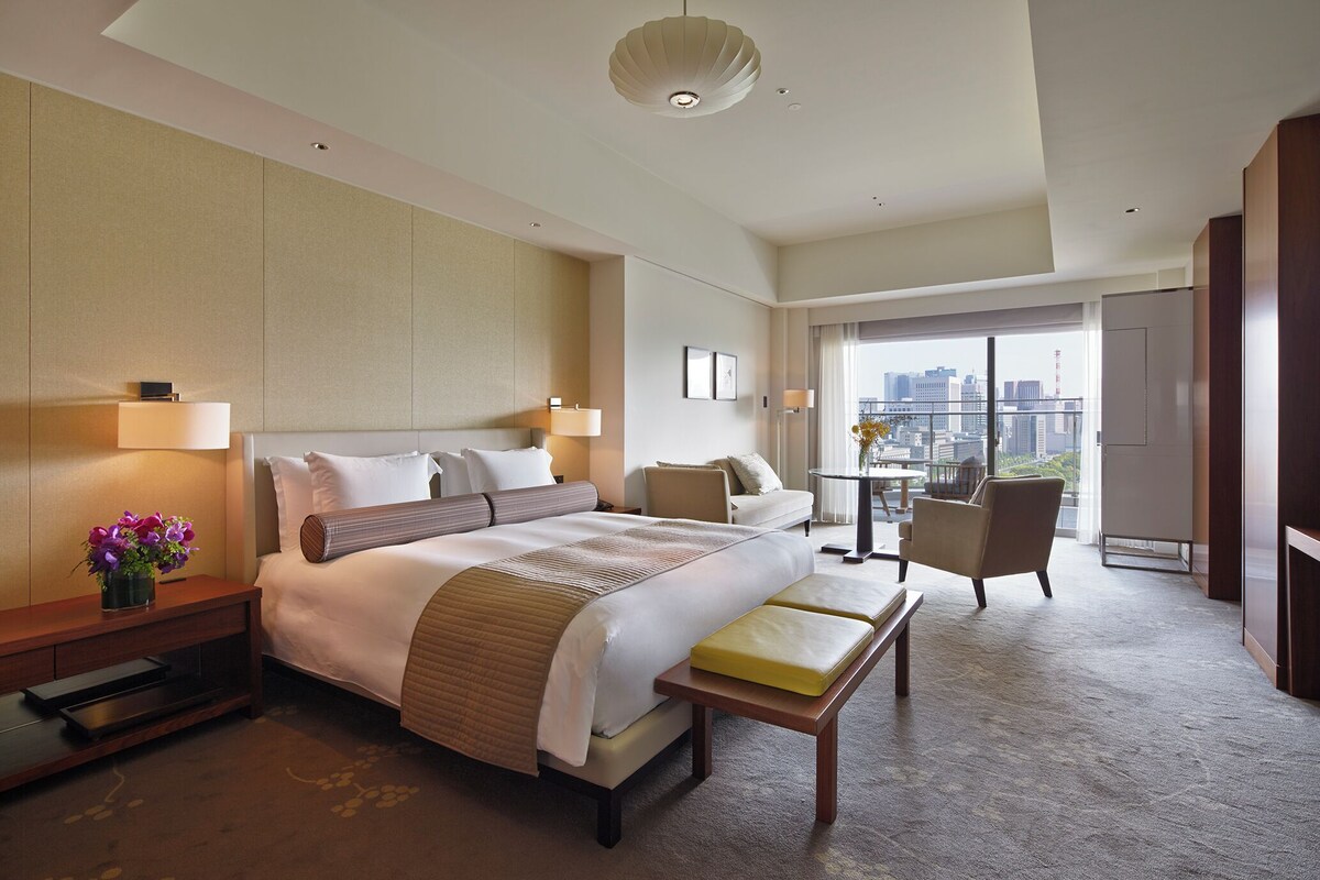 A hotel room with a large bed, bedside tables, a desk, a chair, and a view of the city.