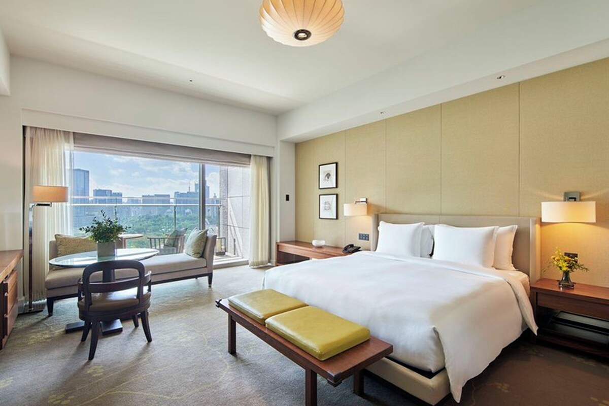 A hotel room with a large bed, a desk with a chair, a small table with a plant, a bench, and a view of the city through large windows.