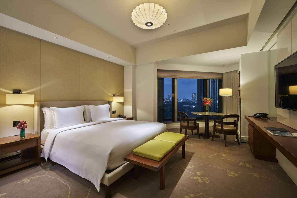 A hotel room with a large bed, a desk, a chair, a television, and a view of the city at night.