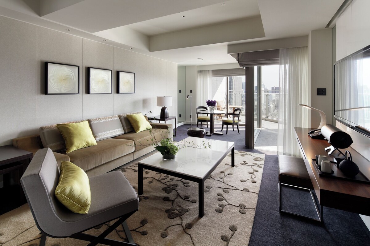 A modern hotel room with a sofa, a coffee table, a desk with a lamp, and a view of the city through large windows.