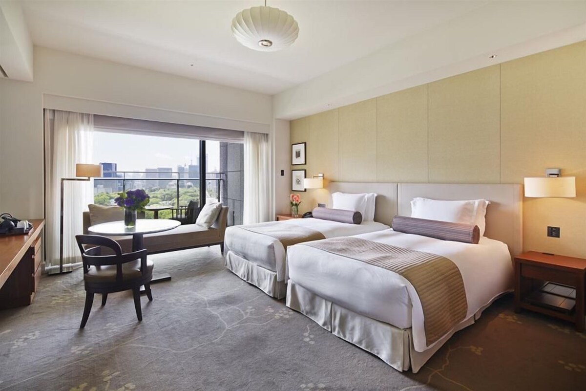 A hotel room with two beds, a desk, a chair, a balcony with a view, and a cityscape visible through the window.
