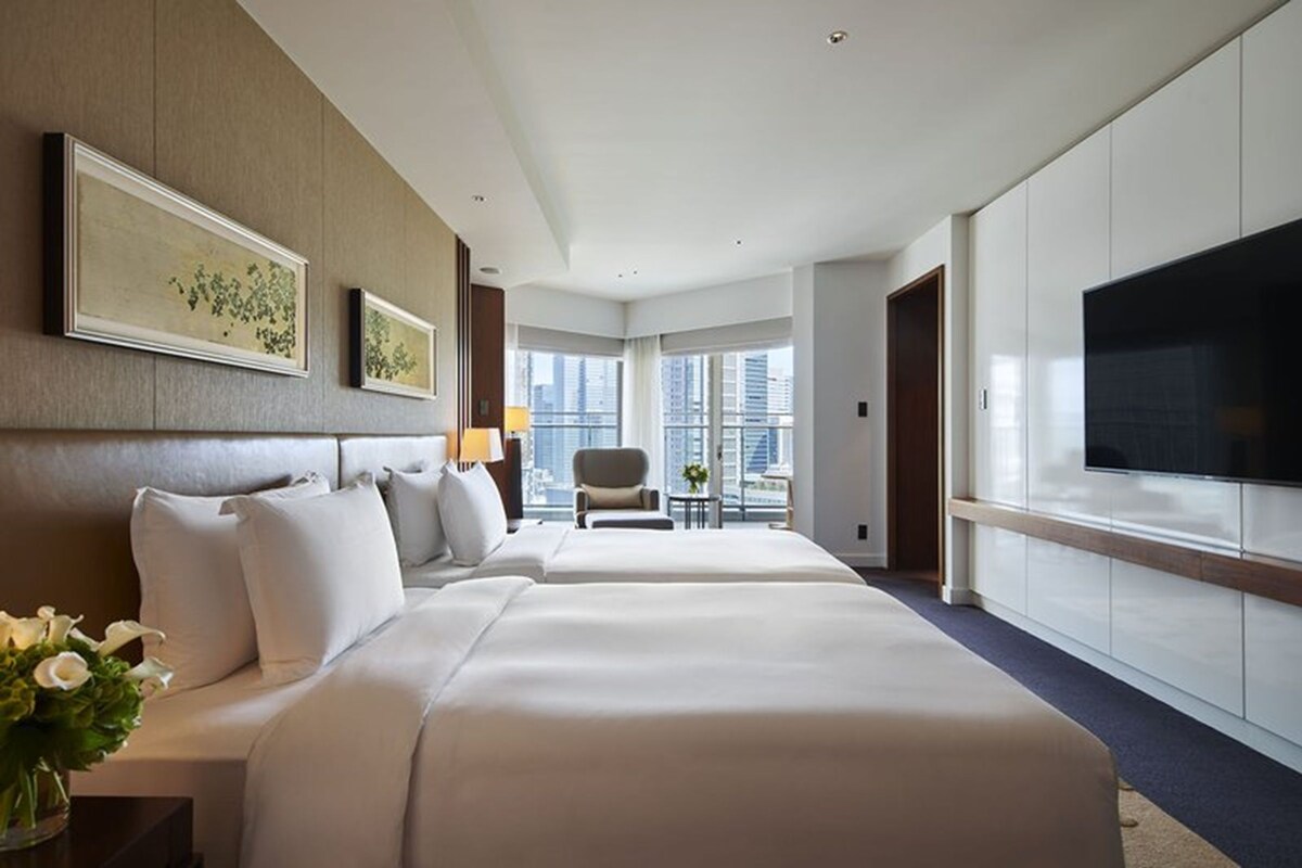 A hotel room with two beds, a TV, a chair, and a view of the city.