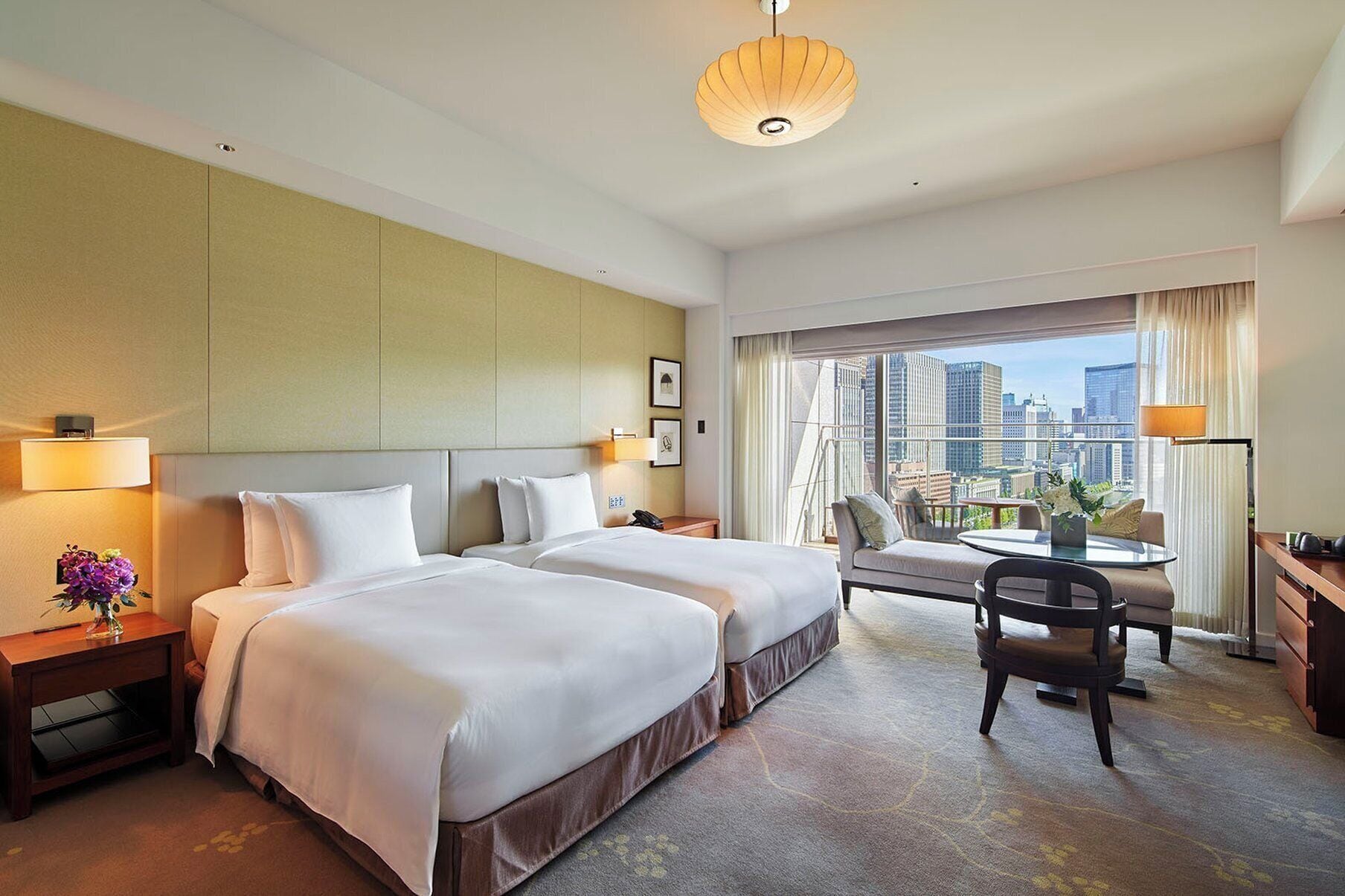 A hotel room with two beds, a desk, a chair, a view of the city, and a large window with curtains.
