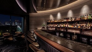 Un bar avec un comptoir incurvé, une grande variété de bouteilles d’alcool et une vue sur la ville la nuit.