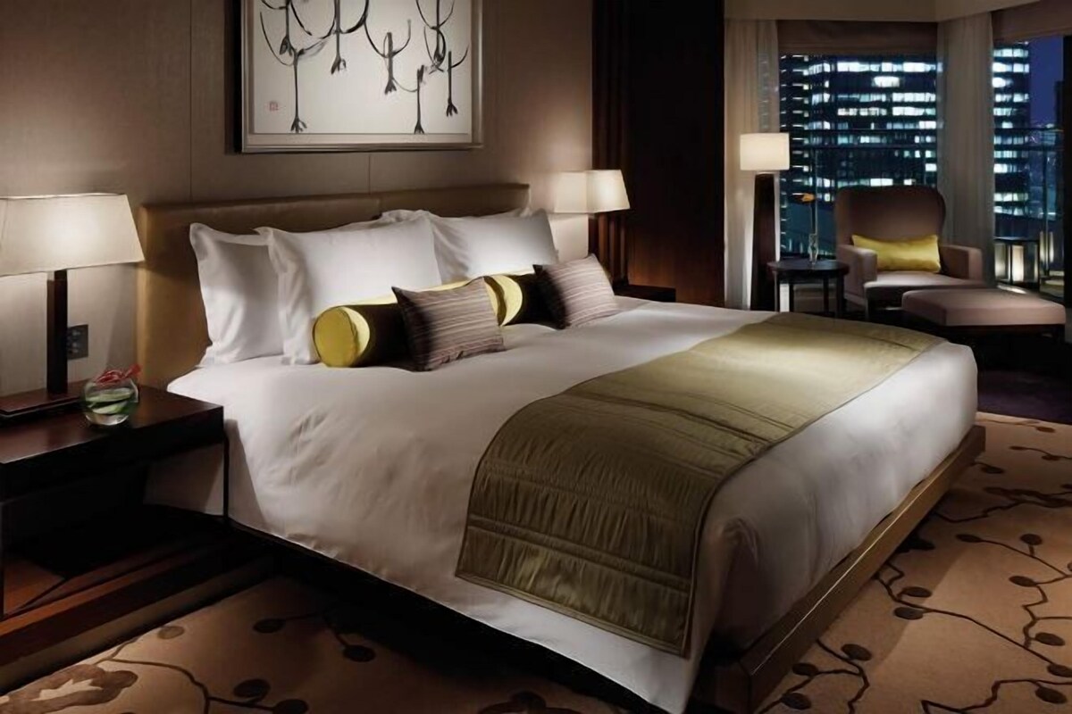 A hotel room with a large bed, two bedside tables, a nightstand, a chair, and a view of the city at night.