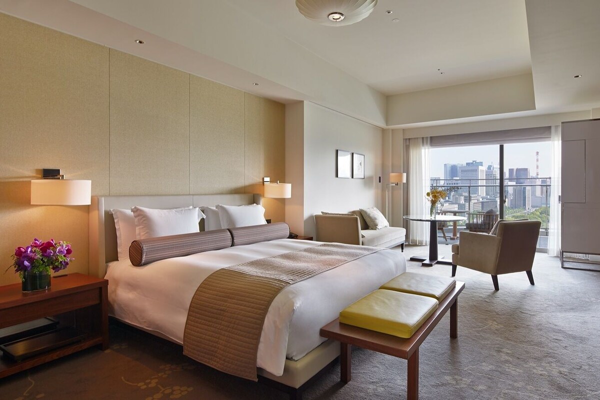 A hotel room with a large bed, a sitting area with a table and chairs, and a balcony with a city view.