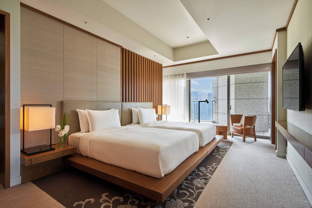 A modern hotel room with two large beds, a flat-screen TV, a balcony with a city view, and a large window.