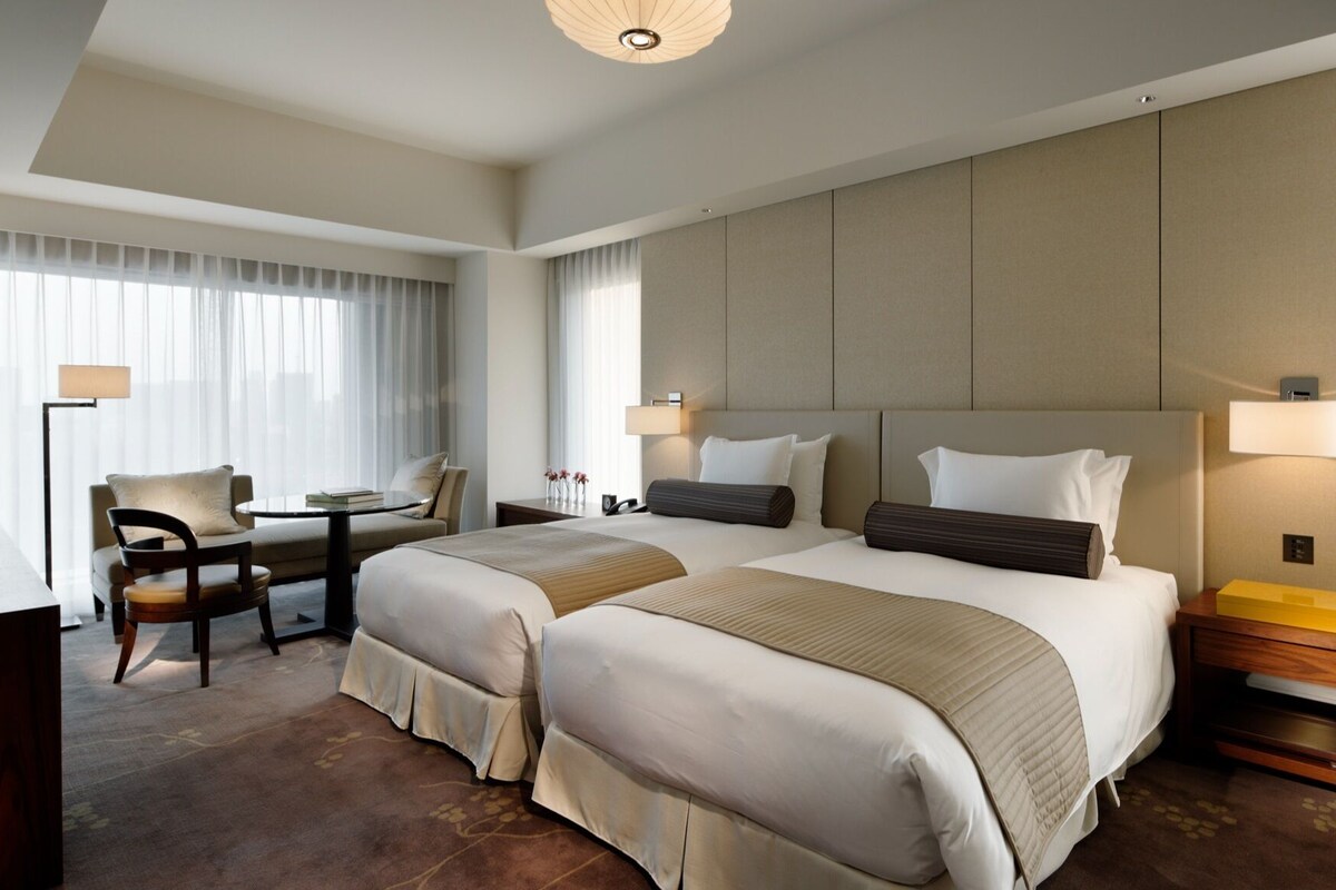 A hotel room with two beds, a desk, a chair, a nightstand, and a lamp.