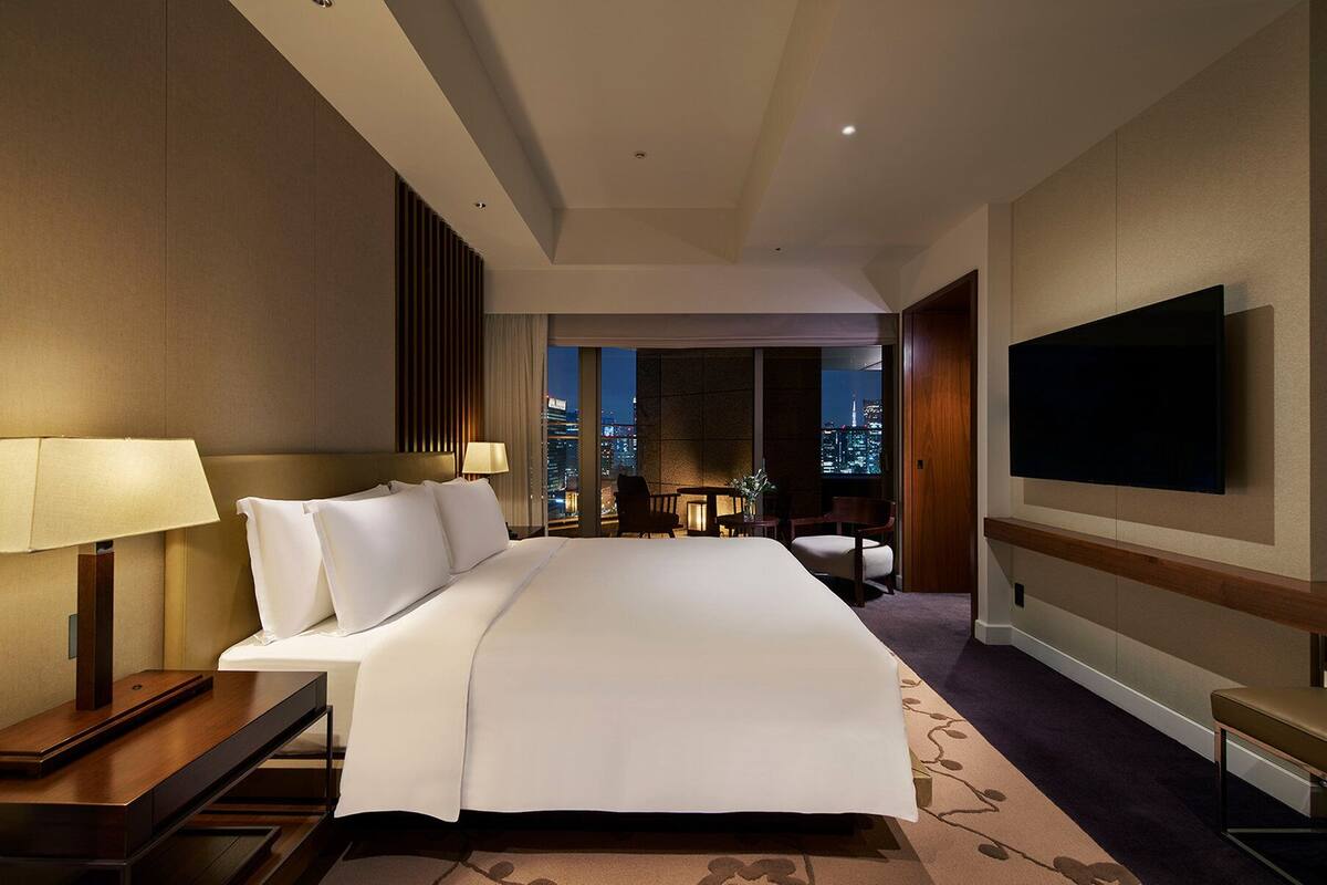A hotel room with a large bed, a desk with a lamp, a TV on the wall, and a view of the city at night.