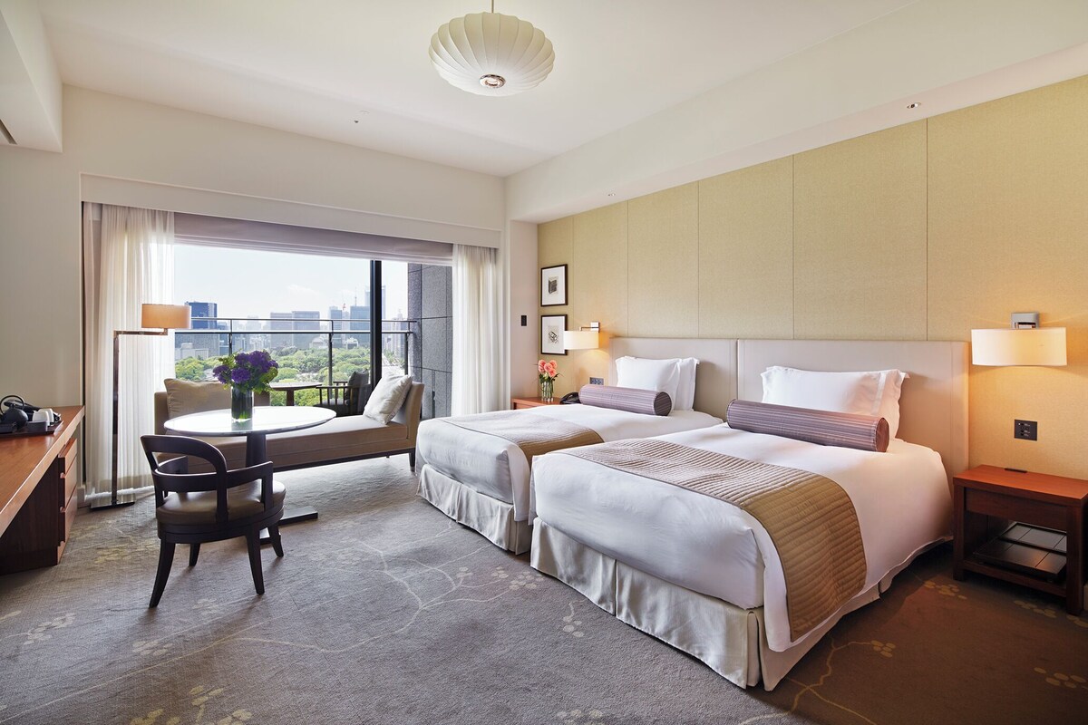 A hotel room with two beds, a desk, a chair, a balcony, and a city view.