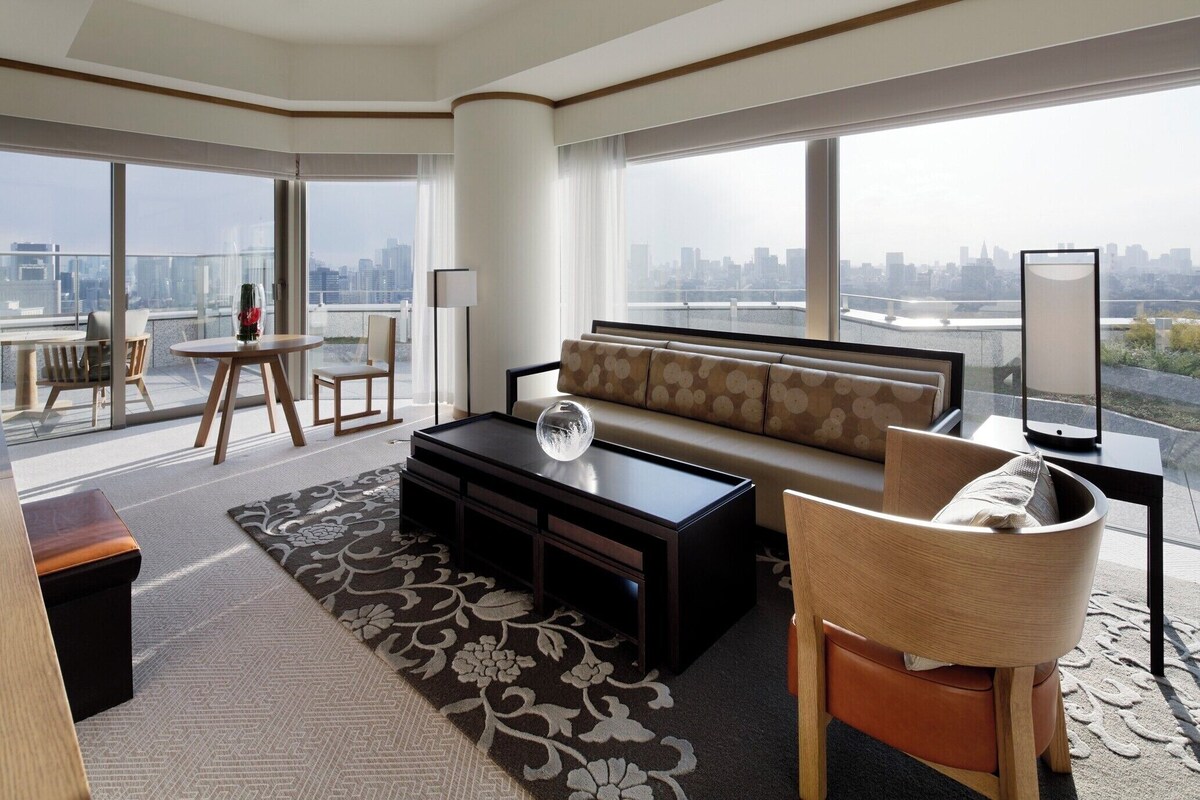 A modern living room with a sofa, coffee table, and chairs, large windows with a city view, and a balcony with a table and chairs.