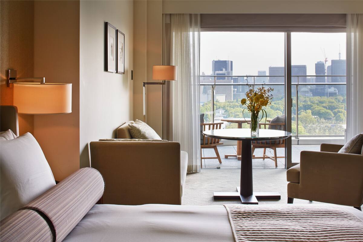 A hotel room with a bed, two armchairs, a dining table with chairs, a lamp, and a view of the city through large windows.