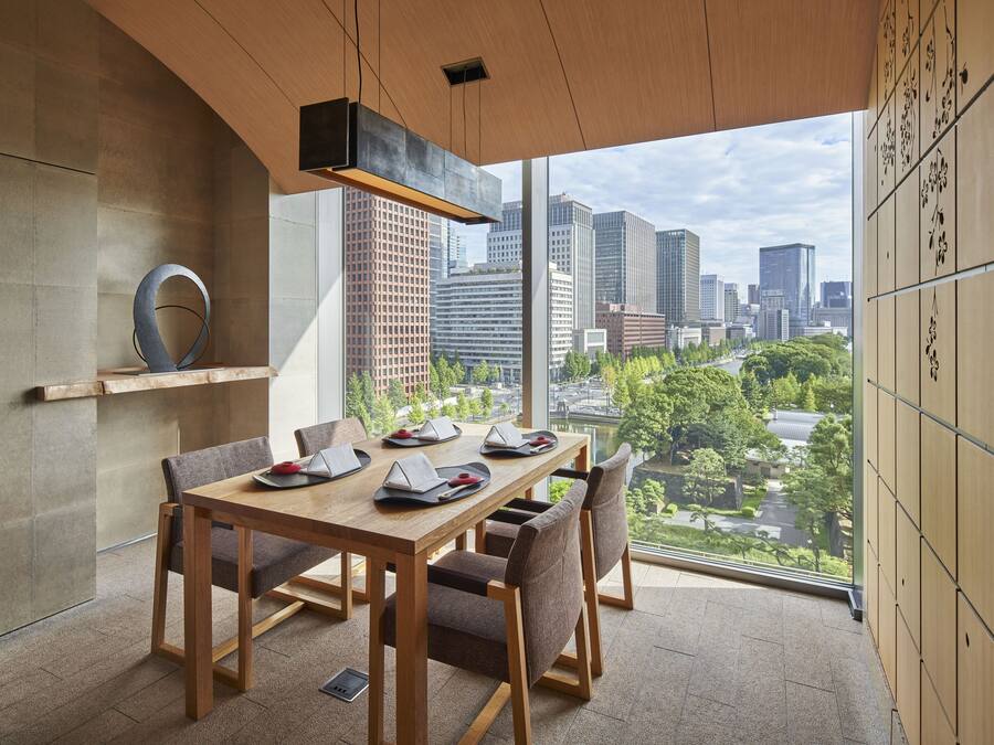 Un comedor moderno con vista a la ciudad, que presenta una mesa y sillas de madera, una lámpara colgante y vistas de edificios altos y vegetación.