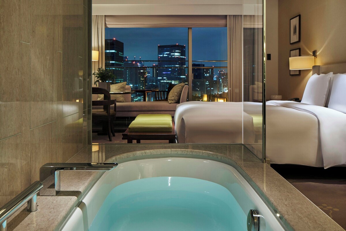 A hotel room with a large bathtub, a bed, a sofa, and a view of a cityscape at night.