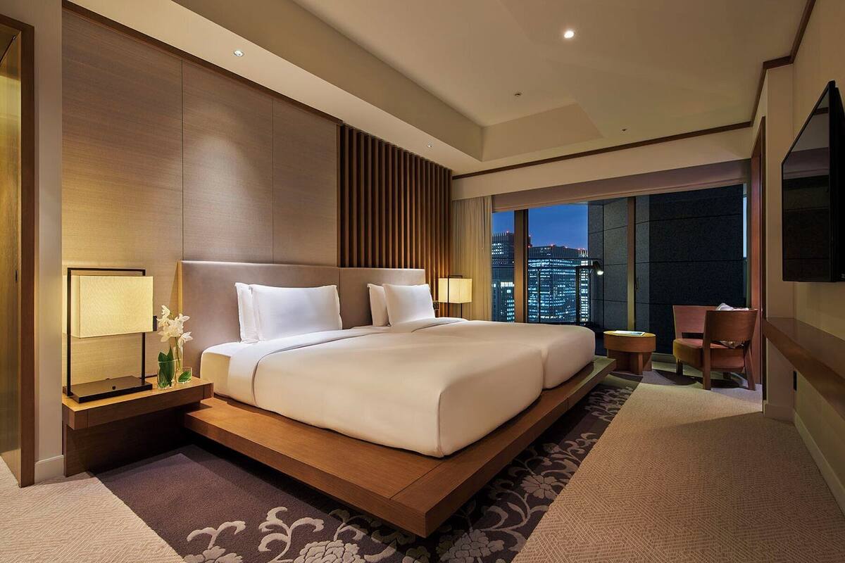 A modern hotel room with a large bed, a city view, and a seating area with a TV.