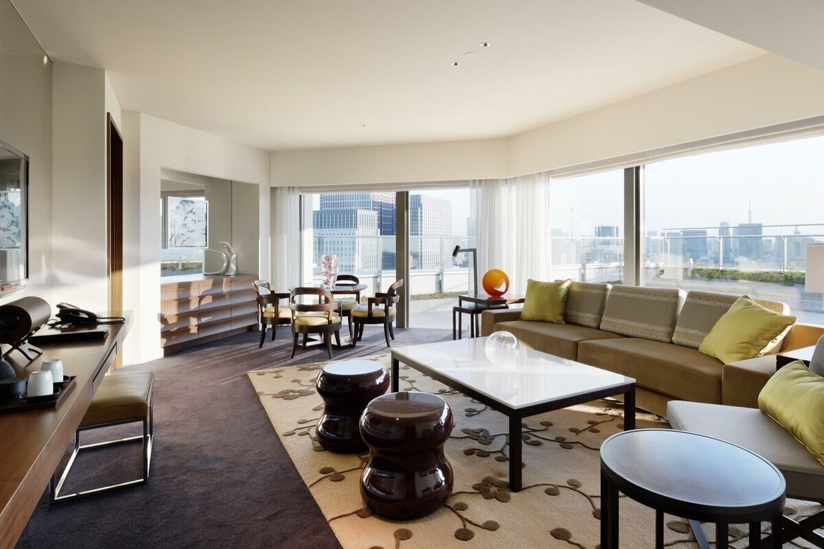 A modern living room with a sofa, coffee table, and chairs, large windows with a city view, and a dining area with a table and chairs.