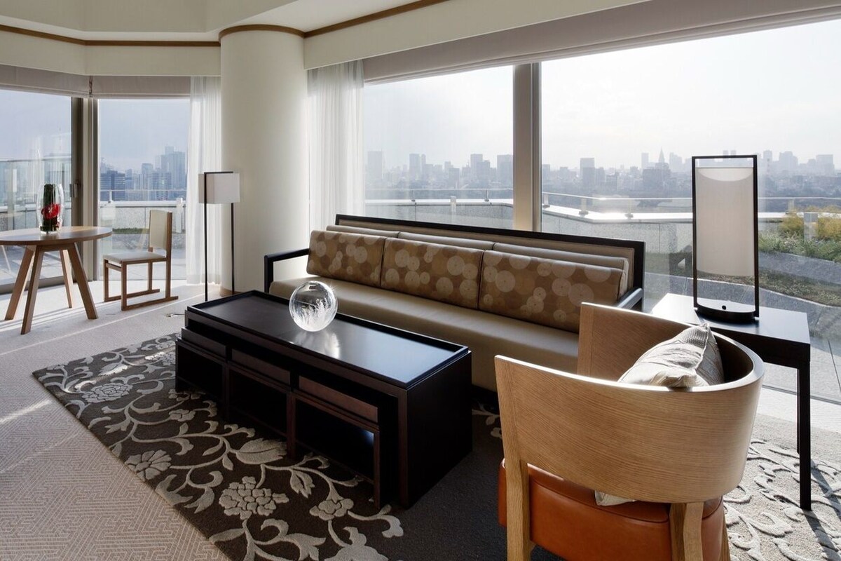 A modern hotel room with a sofa, a coffee table, a dining table, and a view of a city skyline.