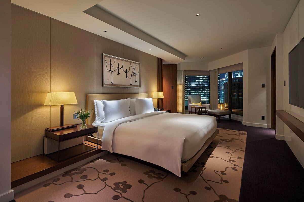 A hotel room with a large bed, two bedside lamps, a nightstand, a chair, and a view of the city at night.