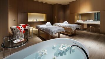 A modern hotel room with a large bathtub, two beds, a sitting area with a chair and table, and a large mirror.