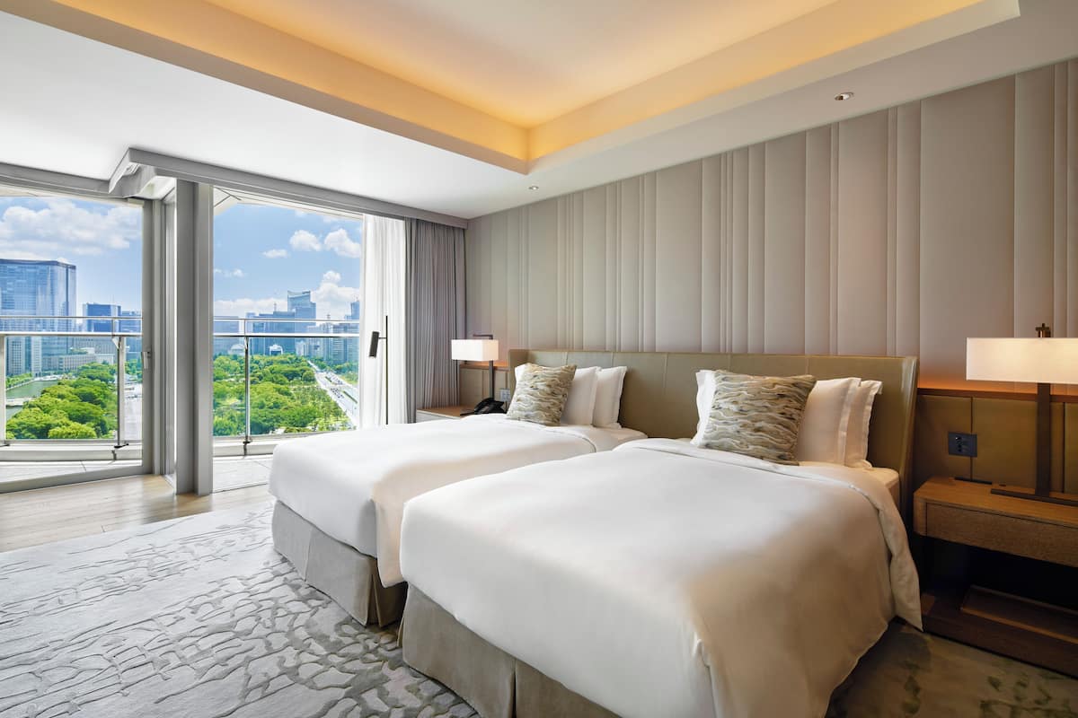 A hotel room with two beds, a large window with a city view, and a balcony.