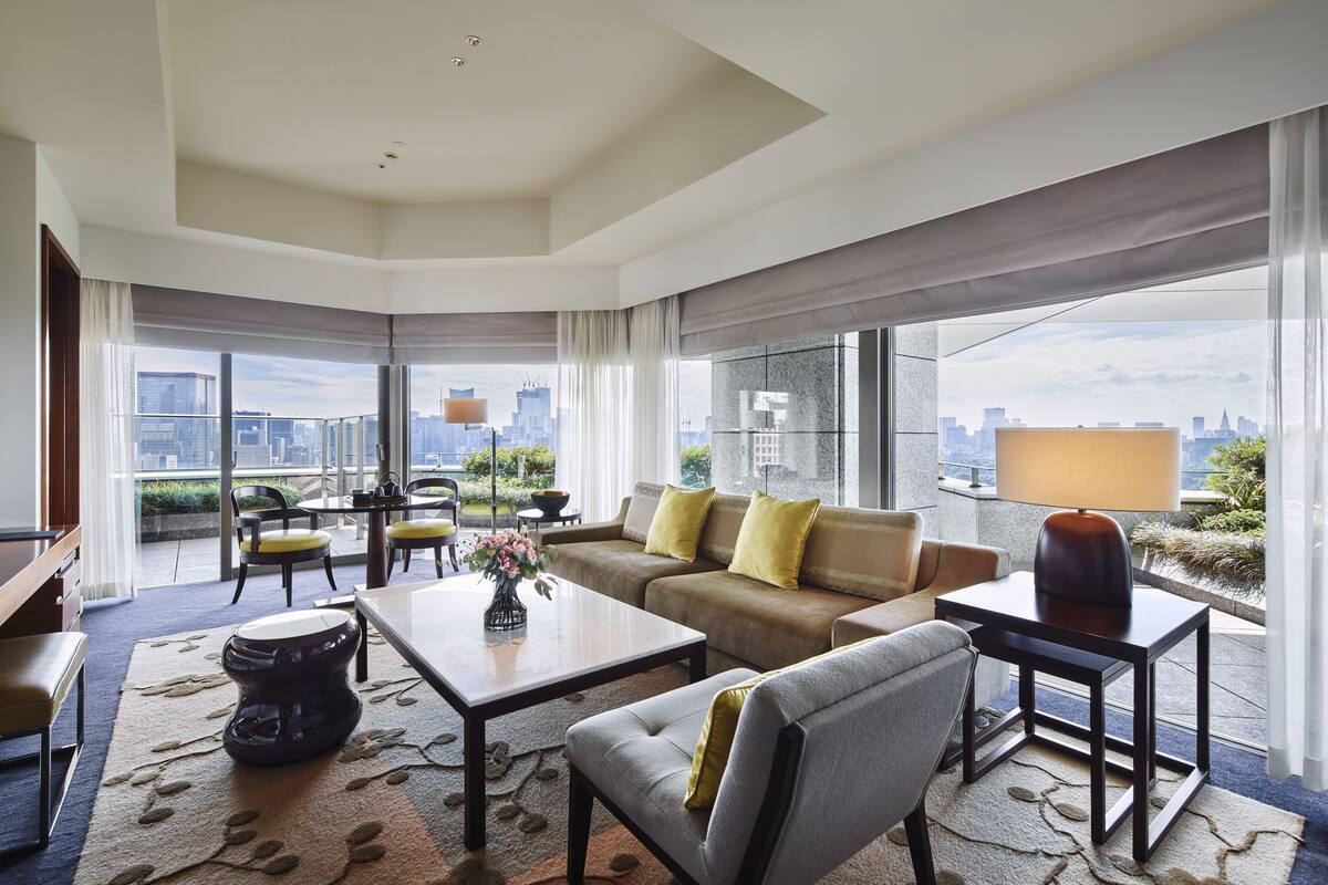 A modern hotel room with a large window, a sofa, a coffee table, and a dining area.