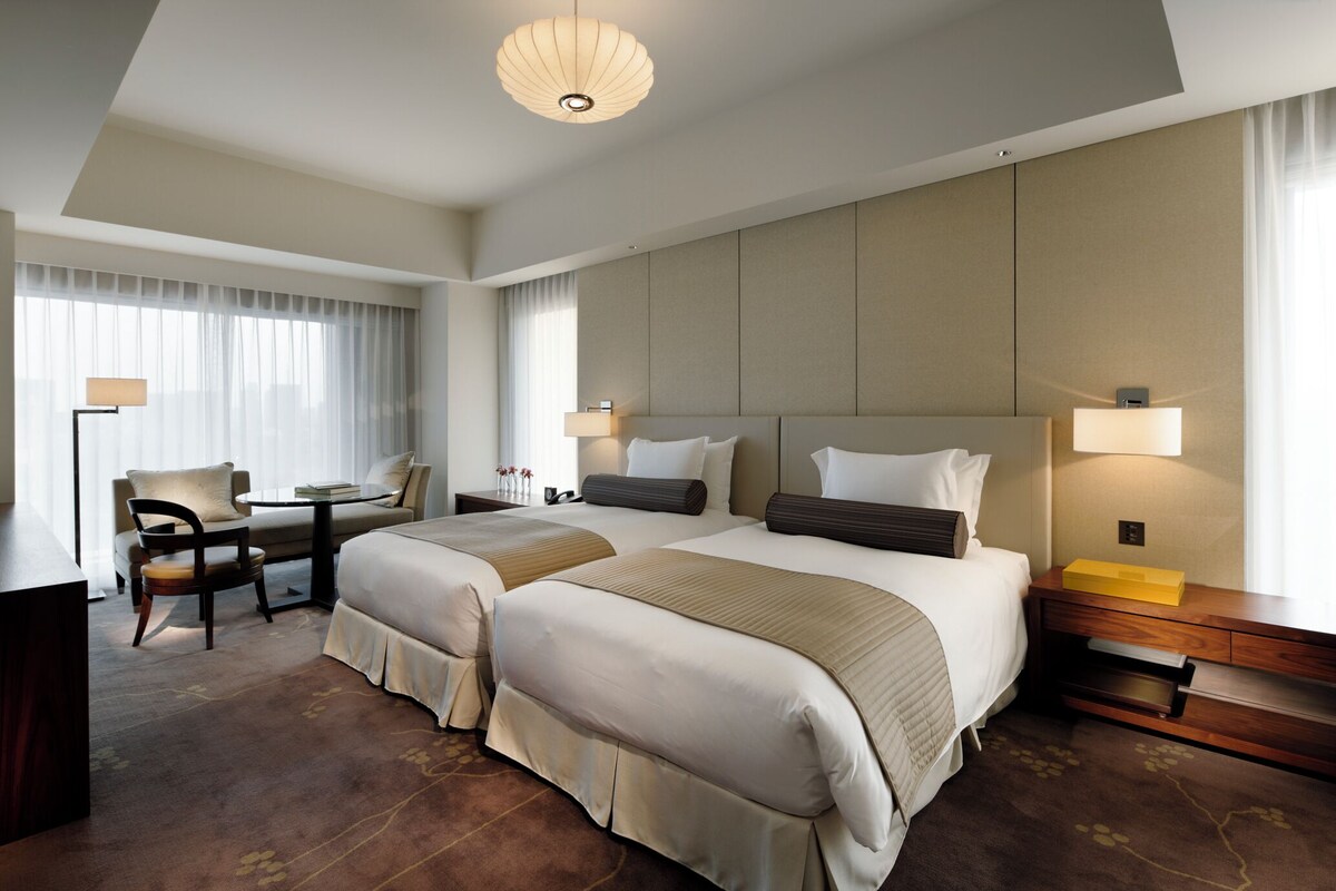 A hotel room with two beds, a desk, a chair, a lamp, and a nightstand.