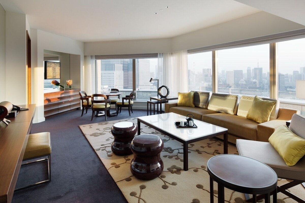 A modern living room with a sofa, coffee table, and chairs, a dining area with a table and chairs, and large windows with a city view.