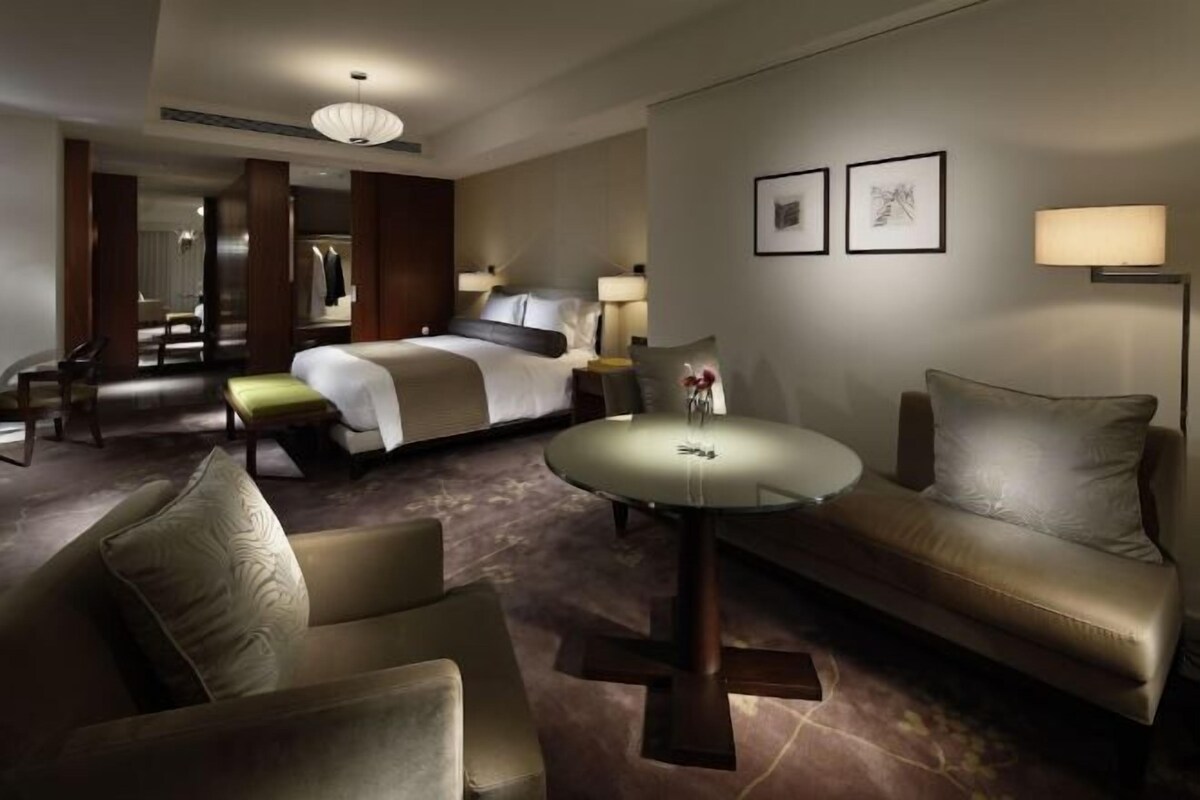 A hotel room with a bed, a sofa, a round table, and a chair. There are two lamps on the wall and a mirror.