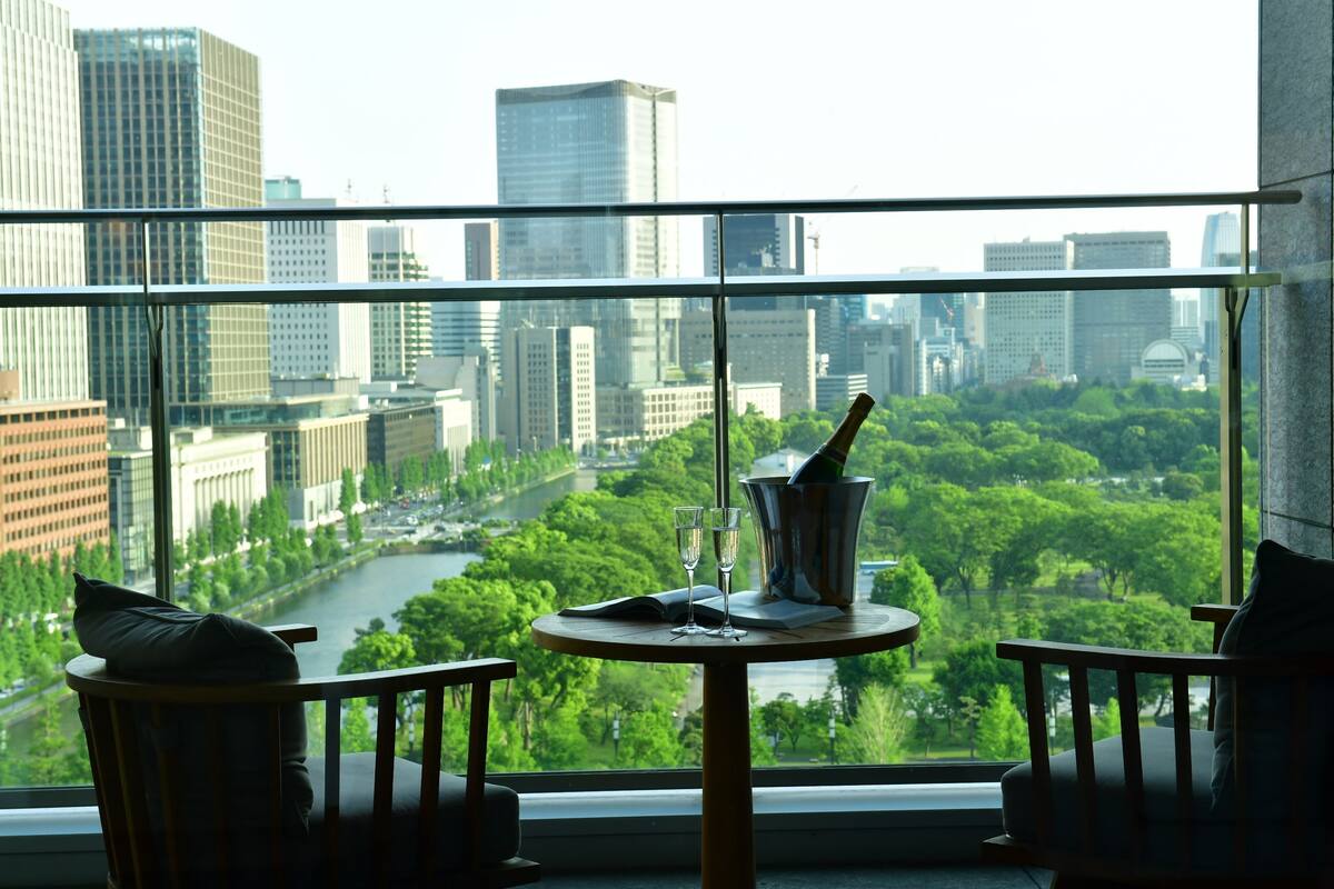 A room with a large window overlooking a cityscape and a river. There is a round table with a bottle, glasses, and a book. Two chairs are positioned in front of the table.