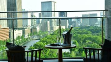 A room with a large window overlooking a cityscape and a river. There is a round table with a bottle, glasses, and a book. Two chairs are positioned in front of the table.