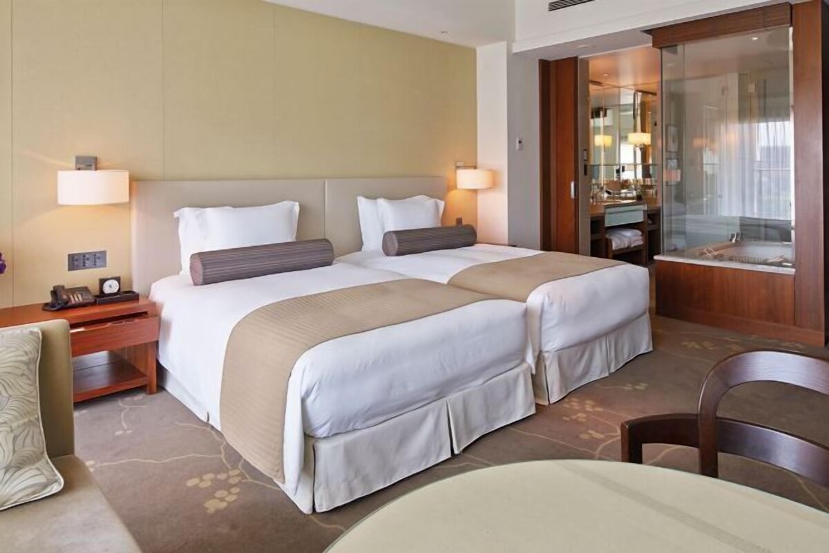 A hotel room with two beds, a nightstand with a lamp, a bathroom with a glass shower enclosure, and a dining area with a table and chair.