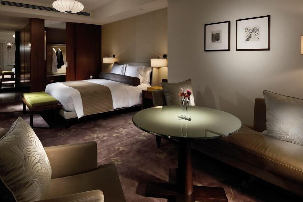 A hotel room with a bed, a round glass table, a sofa, and a chair. There are two framed pictures on the wall.